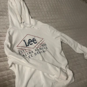Vit hoodie från Lee med tryck - Säljer en vit hoodie från Lee med stort tryck på bröstet i blått och rött. Tröjan har huva med snörning, ribbade muddar och lång ärm. Perfekt för dig som gillar streetwear och vill ha en chill look. Materialet är mjuk bomull som känns skönt mot huden.