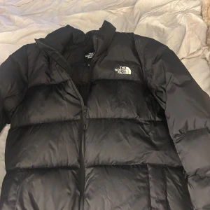 Svart pufferjacka från The North Face - Säljer en svart pufferjacka från The North Face i storlek L. Jackan har ett klassiskt quiltat mönster, hög krage och dragkedja framtill. Loggan är broderad på bröstet. Perfekt för kalla dagar och har en clean, urban vibe.