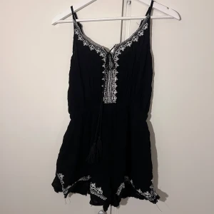 Svarta jumpsuit med broderi från H&C - Svart jumpsuit, köpt i Thailand. Broderade detaljer längs kanterna och vid halsringningen. Det finns snörning framtill vid bröstet, med  tofsar längst ut på snörena.