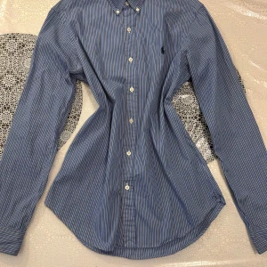 Blåvit randig skjorta Polo Ralph Lauren - Snygg blå skjorta med vita ränder från Polo Ralph Lauren. Modellen är slim fit med klassisk krage och knappar framtill. Materialet är mjuk bomull och skjortan har långa ärmar med manschettknappar. Perfekt för dig som gillar stilrena och tidlösa plagg.