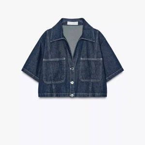 Mörkblå croppad jeansjacka - Snygg croppad jeansjacka i mörkblå denim med korta ärmar, stora bröstfickor och knappar framtill. Jackan har klassisk krage och markerade sömmar för en cool look. Perfekt för dig som vill ha en trendig och avslappnad stil.