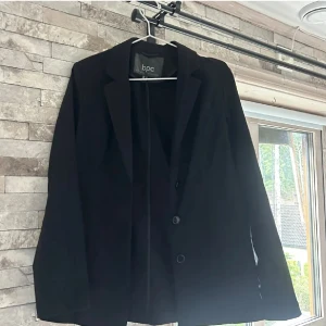 Svart kavaj från bpc, storlek 36 - Stilren svart kavaj från bpc bonprix collection i klassisk modell med två knappar framtill och smala slag. Passar dig som gillar enkel och snygg look. Materialet känns mjukt och har en lätt struktur. Perfekt att matcha med jeans eller kostymbyxor. Har tunn axelvaddning. Använd få gånger 