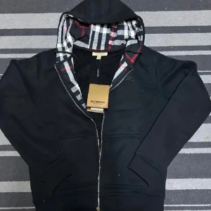 Svart Burberry hoodie - Svart hoodie från Burberry med klassiskt rutig foder i huvan och insidan, dragkedja framtill och broderad Burberry-logga på bröstet. Tröjan har snörning i huvan och två fickor fram. Perfekt för dig som gillar stilren design med lyxig touch.