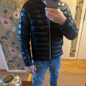 Svart Moncler pufferjacka herr - Snygg svart/mörkblå pufferjacka från Moncler. Inbyggd vind luva som går att vika in. Perfekt till hösten då den är varm och har en stilig look. Materialet är relativt glansigt och  skicket är topp klass. 