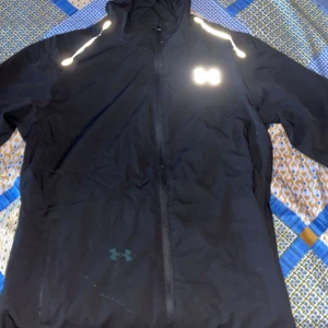 Svart vindjacka från Under Armour - Svart vindjacka från Under Armour med reflekterande detaljer på axlarna och stor logga på ryggen. Jackan har huva, dragkedja framtill och lång ärm. Perfekt för dig som gillar sportig stil och vill synas i mörkret.