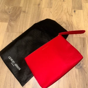 Röd kuvertväska Giorgio Armani - Snygg röd kuvertväska från Giorgio Armani med gulddetaljer och ett coolt prickigt mönster som bildar berg på ena sidan. Väskan har en praktisk handledsrem och dragkedja upptill. Materialet är glansigt och känns lyxigt, perfekt för att lyfta din outfit.