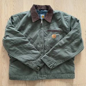 Grön Carhartt workwear jacka M - Snygg grön Carhartt workwear jacka med brunt krage och bruna detaljer vid fickor. Insidan har ett rutigt blått foder. Jackan har dragkedja framtill, långärmad passform och klassisk Carhartt-logga på bröstet. Perfekt för dig som gillar street och utility vibes.