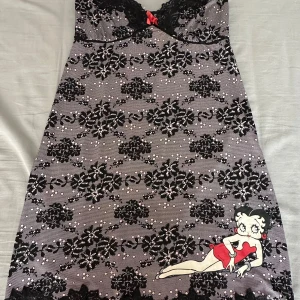 Betty Boop spetsklänning med tryck - Supersöt kort klänning från Betty Boop med svart spets, blommigt mönster och en röd rosett framtill. Klänningen har smala axelband, figurnära passform och en ikonisk Betty Boop-figur tryckt nertill. Materialet är polyester med elastan för extra stretch.