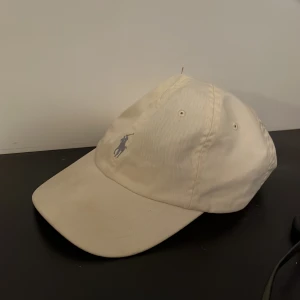 Beige keps från Polo Ralph Lauren - Snygg beige keps från Polo Ralph Lauren med klassisk broderad logga framtill och justerbart spänne bak. Kepsen är i bomull och har ventilerande hål på ovansidan. Perfekt accessoar för en clean och stilren look. 