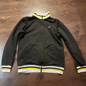 Svart ziptröja Norra Ståplats M - Svart ziptröja från 2002 Produktion med Norra Ståplats-märke på bröstet. Tröjan har ribbade muddar och krage i grått, vitt och gult, samt hel dragkedja framtill och två fickor. Perfekt för dig som gillar sportig stil och snygga detaljer. Pris kan diskuteras 