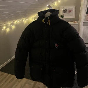 Svart dunjacka Fjällräven XS - Svart dunjacka från Fjällräven i storlek XS. Jackan har en huva med gula snören och klassisk Fjällräven-logga på bröstet. Stängs med både dragkedja och knappar. Perfekt för kalla vinterdagar.