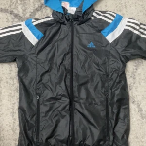 Svart vindjacka från Adidas med blå och vita detaljer - Svart vindjacka från Adidas med blå och vita detaljer samt klassiska vita ränder längs ärmarna. Jackan har huva, dragkedja framtill och två fickor med dragkedja. Perfekt för dig som gillar sportig stil och vill ha något lätt och smidigt.