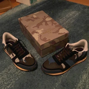 Valentino Garavani Rockrunner camo sneakers - Säljer ett par Valentino Garavani Rockrunner sneakers i storlek 43. Skorna har ett snyggt kamouflagemönster i blått, svart och vitt med svarta snören och detaljer. Ovandelen är i mesh och mocka, med en klassisk gummisula. Kommer med originalkartong.