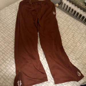 Flared Bruna mjukisbyxor  - Bruna mjukisbyxor med vita broderade loggor på höften och vid bensluten. Byxorna har dragkedjor längst ner på benen och resår i midjan med snörning. Perfekta för en chill och avslappnad stil.