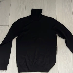 Svart polotröja från J.Lindeberg - Svart polotröja från J.Lindeberg i klassisk modell med lång ärm och hög krage. Tröjan är stilren och passar perfekt till lager-på-lager. Tillverkad i mjukt material som känns skönt mot huden.