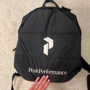 Svart ryggsäck från Peak Performance - Svart ryggsäck från Peak Performance med stor vit logga och text på framsidan. Väskan har en rundad form, justerbara axelremmar och ett praktiskt handtag upptill. Perfekt för skola eller träning och rymmer allt du behöver under dagen.