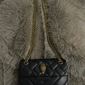 Svart Kurt geiger axelväska med kedja - Säljer en svart Kurt axelväska med guldfärgad kedjerem och unik handformad detalj på locket. Väskan är i skinn och har en lyxig känsla, perfekt för dig som vill sticka ut med accessoarerna.