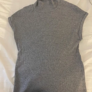 Grå ribbad polotopp - Snygg grå polotopp med korta ärmar. Toppen har en hög krage och är tillverkad i ett mjukt, stretchigt material som sitter skönt mot kroppen. Storlek M men passar även S.