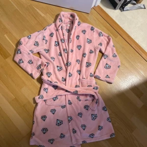 Rosa morgonrock med diamantmönster - Mysig rosa fleecejacka med huva och bälte i midjan. Jackan har ett coolt diamantmönster över hela och två praktiska fickor framtill. Perfekt för att chilla hemma eller mysa runt i. Långärmad och supermjuk känsla.