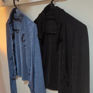 Set 2 st: Tröjor med knappar Cropped Toppar Stl M - Rynkad (Svart & Blå) - Hög Krage/Polo Dam Stl M - Säljer ett snyggt paket med två matchande, långärmade toppar i en trendig cropped-modell (kort modell). Båda topparna har en uttrycksfull design med rynkning/drapering längs framsidan, vilket skapar en smickrande textur. De har en hög, polo-inspirerad krage och stängs med en knappslå. Perfekt för utekvällar eller en cool lager-på-lager-look.