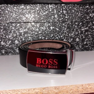 Hugo boss bälte  - Snyggt svart bälte från Hugo Boss med stilren, blank metallspänne där BOSS-loggan syns tydligt i rött och guld. Bältet är gjort i slätt skinn och har en klassisk, rak form som passar till jeans eller kostymbyxor. Perfekt accessoar för att lyfta din outfit.