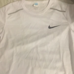 Vit Nike t-shirt i funktionsmaterial - Vit t-shirt från Nike i funktionsmaterial, perfekt för träning. Klassisk rund halsringning, korta ärmar och svart Nike-logga på bröstet. Lätt och luftig känsla med diskreta grå detaljer vid axlarna. Storlek M men jag skulle säga passar som s eller xs