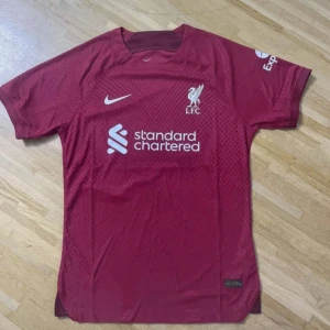 Liverpool FC vinröd Nike fotbollströja - Liverpool FC matchtröja i vinrött med korta ärmar och rund hals. Nike-logga och Liverpool-märke på bröstet, samt Standard Chartered tryck. Tillverkad i lätt, ventilerande polyester med subtilt geometriskt mönster. Perfekt för dig som vill representera LFC på träning eller match.