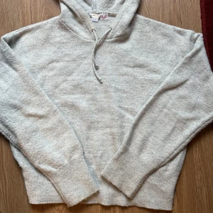 Ljusgrå stickad hoodie från Gina Tricot - Mysig ljusgrå stickad hoodie från Gina Tricot med huva och snörning. Tröjan har långa ärmar och breda ribbade muddar vid ärmslut och nederkant. Perfekt för kyliga dagar när du vill vara både bekväm och snygg.