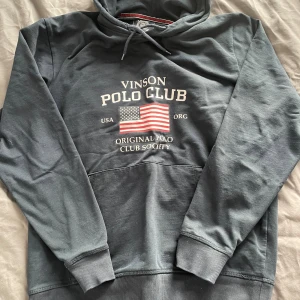 Mörkblå hoodie från Vinson Polo Club - Snygg mörkblå hoodie från Vinson Polo Club med tryck av amerikanska flaggan och text på bröstet. Tröjan har huva med snörning och en klassisk känguruficka framtill. Perfekt för en chill och sportig stil.