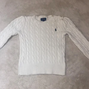 Vit kabelstickad tröja Polo Ralph Lauren - Vit kabelstickad tröja från Polo Ralph Lauren med rund halsringning och ribbade muddar. Klassisk marinblå logga broderad på bröstet och knappar på axeln. Perfekt för lager-på-lager under höst och vinter.