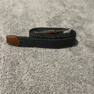 Svart och vit flätad bälte - Snyggt svart och vitt flätat bälte med bruna skinn-detaljer vid spännet och änden. Bältet har ett diskret mönster och ett klassiskt metallspänne. Perfekt accessoar för att lyfta din outfit med en stilren touch.