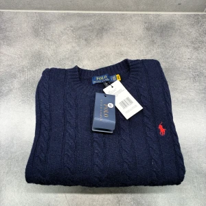 Mörkblå kabelstickad tröja Polo Ralph Lauren - Kabelstickad mörkblå tröja från Polo Ralph Lauren med klassisk röd broderad logga på bröstet. Tröjan har rund halsringning, ribbade muddar och är tillverkad i mjuk ull. Perfekt för dig som gillar en tidlös och stilren look.