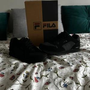 Svarta sneakers från FILA, storlek 42 (använd 1 gång - Säljer ett par svarta FILA sneakers i storlek 42. Skorna har en snygg mix av syntet och textil med diskret mönstrad panel på sidorna. Klassisk snörning och platt sula gör dem perfekta för en clean streetstil. Passar dig som gillar stilrena och bekväma sneakers. Använda en gång så inga skador