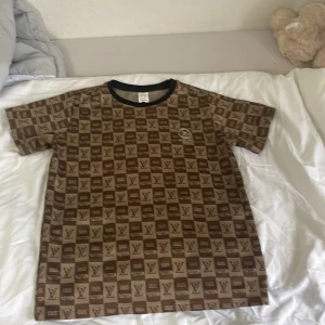 Brun Louis Vuitton-inspirerad t-shirt - Brun t-shirt med korta ärmar och rutigt LV-mönster över hela plagget. Svart ribbad krage och tryckt Louis Vuitton-logga framtill. Skön och avslappnad passform, perfekt för dig som gillar statement-plagg och streetstyle.