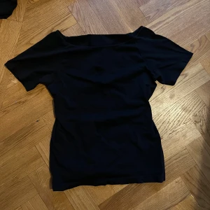 Svart offshoulder topp i bomull - Snygg svart topp med offshoulder-design och korta ärmar. Modellen är figurnära och har en enkel, stilren look utan mönster eller detaljer. Tillverkad i mjuk bomull som känns skön mot huden. Perfekt för dig som gillar minimalistisk stil och vill ha en basic topp i garderoben.