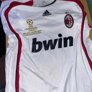 AC Milan Kaka Champions League tröja - Säljer en officiell AC Milan fotbollströja från Champions League-finalen 2007 med Kaka och nummer 22 på ryggen. Tröjan är vit med röda och svarta detaljer, broderat guldmärke och klubbemblem. Tillverkad i lätt polyester, perfekt för fans och samlare.