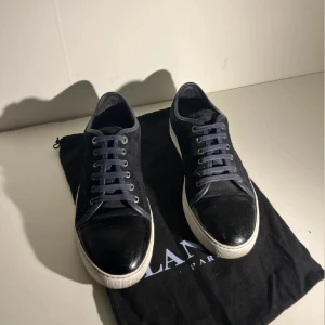 Svarta sneakers från lanvin - Snygga svarta sneakers från lanvin vit platt sula och snörning. Ovandelen är i mocka med detaljer i blankt skinn på tån. Klassisk rund tå och stilren design som passar till streetwear. Kommer med original dustbag. Uk8