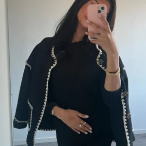 Svart cardigan jacka med vita detaljer - Stilren svart cardigan jacka med vita, sicksack-formade kanter och dekorativa knappar. Jackan har två fickor framtill och är långärmad med en rak, loose passform. Perfekt för dig som gillar en klassisk men trendig look. Ursprungligt pris 700