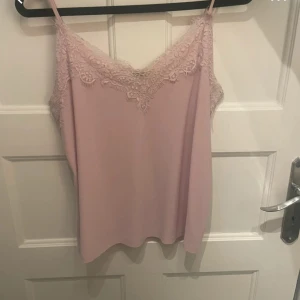 Ljusrosa linnetopp med spets från Amisu - Supersöt ljusrosa linnetopp från Amisu med tunna axelband och vacker spetsdetalj längs urringningen. Toppen har en mjuk, flowy passform och är perfekt för en fräsch och feminin look. Materialet känns lätt och svalt mot huden.