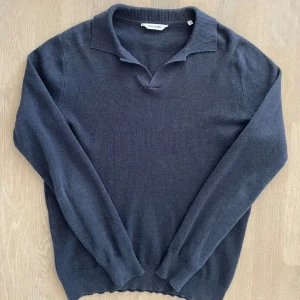 Marinblå bomullströja från John Henric - Säljer en stilren mörkgrå långärmad tröja från John Henric i 100% bomull. Tröjan har en klassisk krage med öppning och ribbade muddar vid ärmslut och nederkant. Perfekt för dig som gillar en clean och enkel look.