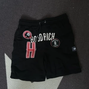 Svarta Hoodrich shorts med patchar - Svarta shorts från Hoodrich med feta broderade patchar och loggor framtill. Snygg snörning i midjan och coola detaljer som sticker ut. Materialet känns mjukt och skönt, perfekt för chill eller streetwear. Hoodrich-etikett på sidan och tryck även baktill.