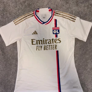 Vit Olympique Lyonnais fotbollströja Adidas - Snygg vit Olympique Lyonnais matchtröja från Adidas med guldiga detaljer och logga. Tröjan har korta ärmar, klassiska Adidas-ränder på axlarna och en röd-blå rand längs framsidan. Tillverkad i lätt och ventilerande material, perfekt för fotboll eller träning.