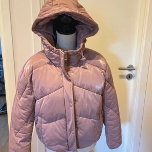 Rosa pufferjacka med huva - Säljer en snygg rosa pufferjacka med stor huva och dragkedja framtill. Jackan är quiltad och har tryckknappar samt snörning vid huvan. Perfekt för kalla dagar när du vill vara både varm och trendig.