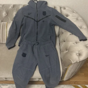 Grå Nike FC Barcelona hoodie M - en mörkblå zip up hoodie från Nike med FC Barcelona-logga och Spotify-tryck. Tröjan har hel dragkedja, huva och långa ärmar. Perfekt för dig som älskar fotboll och vill ha en sportig stil. Materialet är mjukt och skönt, passar till chill eller träning.