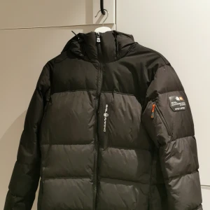 Svart pufferjacka Sail Racing S - Svart pufferjacka från Sail Racing med luva och dragkedja. Jackan har patch på ärmen med 'Sail Antarctica Expedition' och logga på bröstet. Fylld för extra värme och perfekt för kalla dagar. Snygg, sportig look och praktiska fickor. Den är köpt ungefär för ett år sen. I storlek S. 