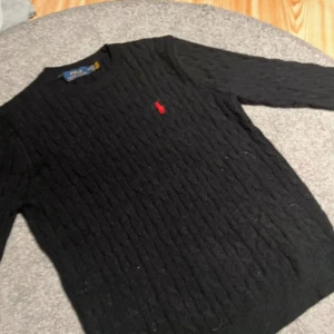 Svart kabelstickad tröja Polo Ralph Lauren - Svart kabelstickad tröja från Polo Ralph Lauren i storlek XXL men passar bra på mig och jag är large/medium (som ni ser på bild 3). Klassisk modell med rund hals och röd broderad logga på bröstet. Tröjan har långa ärmar och ribbade muddar. Perfekt för dig som gillar stilren och tidlös design.