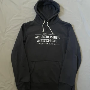 Svart hoodie Abercrombie & Fitch | M - Svart hoodie från Abercrombie & Fitch med vit logga framtill och vita snören i huvan. Tröjan är lite nopprig men inget som påverkar helheten! 