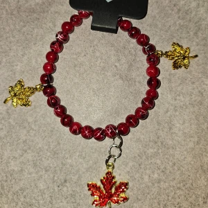 Rött handgjort armband med lönnlöv - Unikt armband med röda pärlor med mönster, dekorerat med tre hängande lönnlöv i guld och rött. Löven ger en höstig vibe och pärlorna har en cool marmorerad look. Perfekt accessoar för dig som vill sticka ut med detaljer. Ca 16cm, gjort av 4mm glaspärlor 