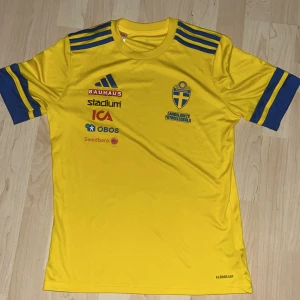 Svenska landslagets fotbollströja Adidas - Gul fotbollströja från Adidas med blå detaljer och tryck från Svenska landslaget. Tröjan har korta ärmar, rund hals och flera sponsorloggor på bröstet. Materialet är lätt och andas, perfekt för träning eller match. Klassisk landslagslook med blå ränder på axlar och ärmar. ÄR EN GÅVA FRÅN SVENSKA FOTOBOLLSFÖRBUNDET SÅ DEN ÄR ÄKTA