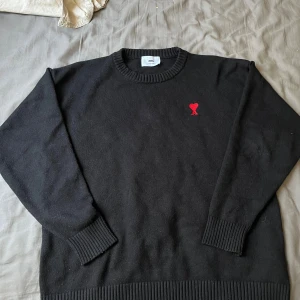 Svart stickad tröja från Ami Paris - Svart stickad tröja från Ami Paris med rund halsringning och ribbade muddar. På bröstet finns Ami:s ikoniska röda hjärta med A-broderi. Replika. Perfekt för dig som gillar stilrena och trendiga plagg med en twist.